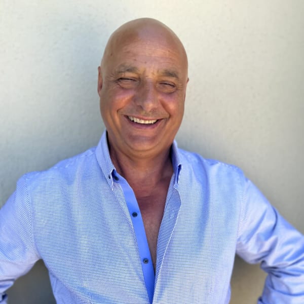A smiling, bald man in a blue button-down shirt stands against a light wall.