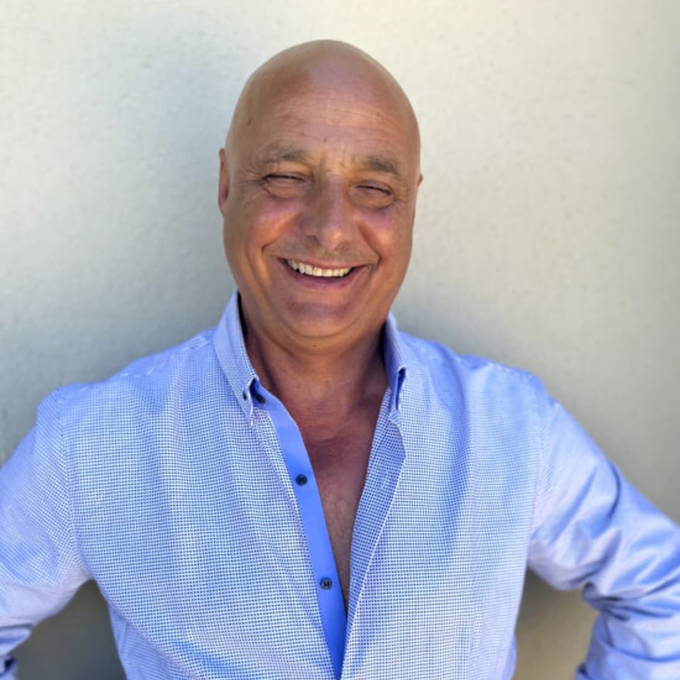 A smiling, bald man in a blue button-down shirt stands against a light wall.