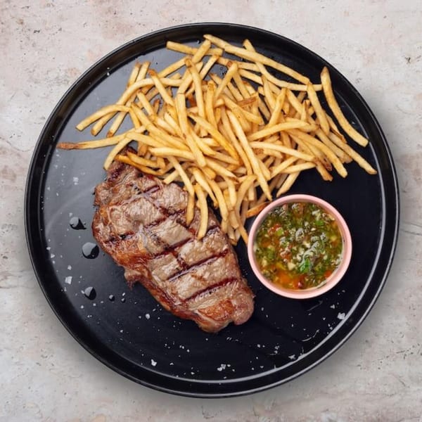Grilled steak with grill marks, a pile of golden french fries, and a small bowl of green sauce.
