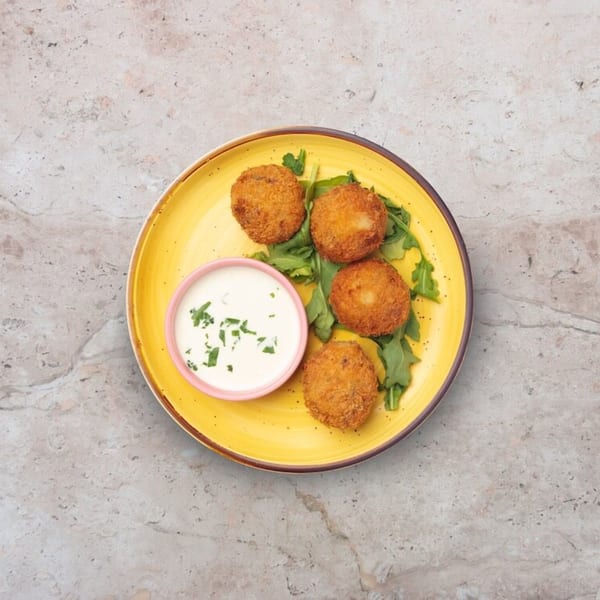 Four golden-brown arancini balls on a yellow plate with arugula and a side of white dipping sauce.