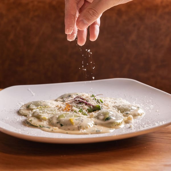 A hand sprinkles grated Parmesan cheese over a plate of Nardò Italian ravioli in cream sauce.