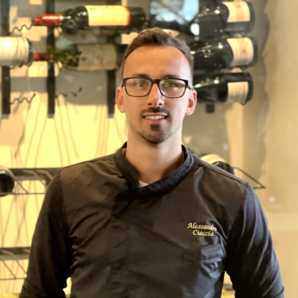 Alessandro Ciaccia, a man in a black chef's jacket, smiles in front of a wine rack.