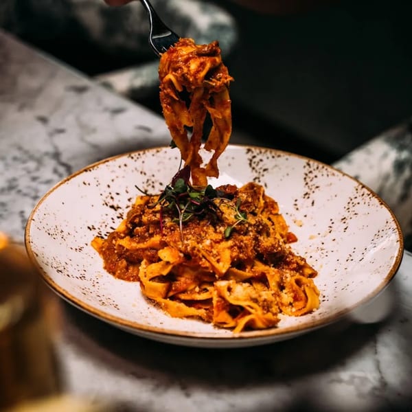 A fork lifts tagliatelle pasta with rich red sauce and microgreens from a white plate.