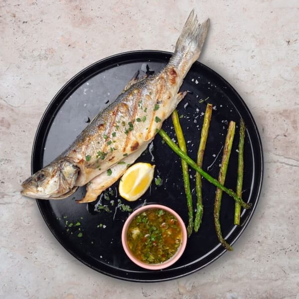 Grilled whole fish with lemon, asparagus, and a side of green sauce on a black plate.